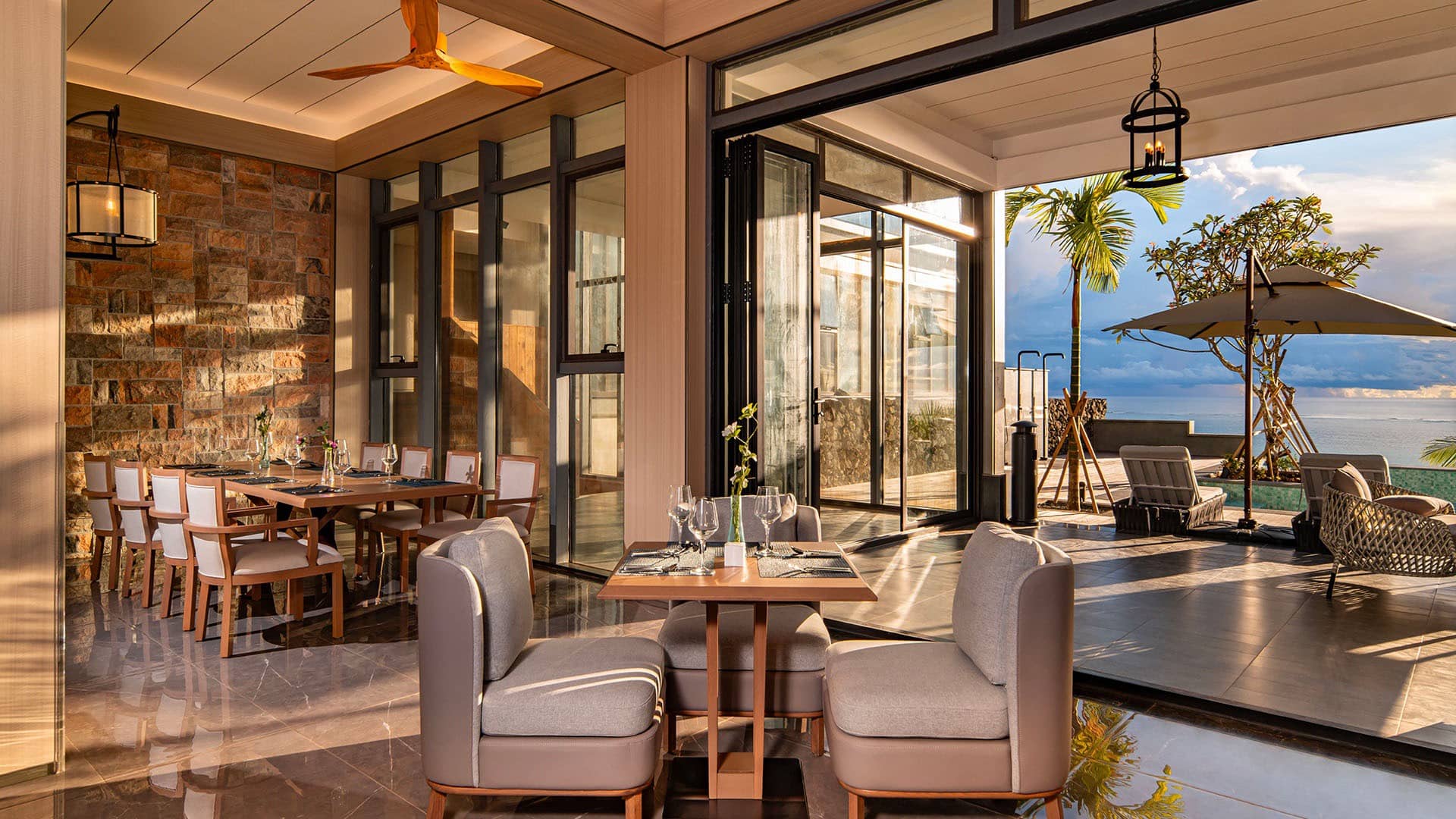 Modern restaurant interior with exposed brick wall, glass wine cellar, wooden dining tables, beige armchairs, and open doors leading to terrace with ocean views at sunset