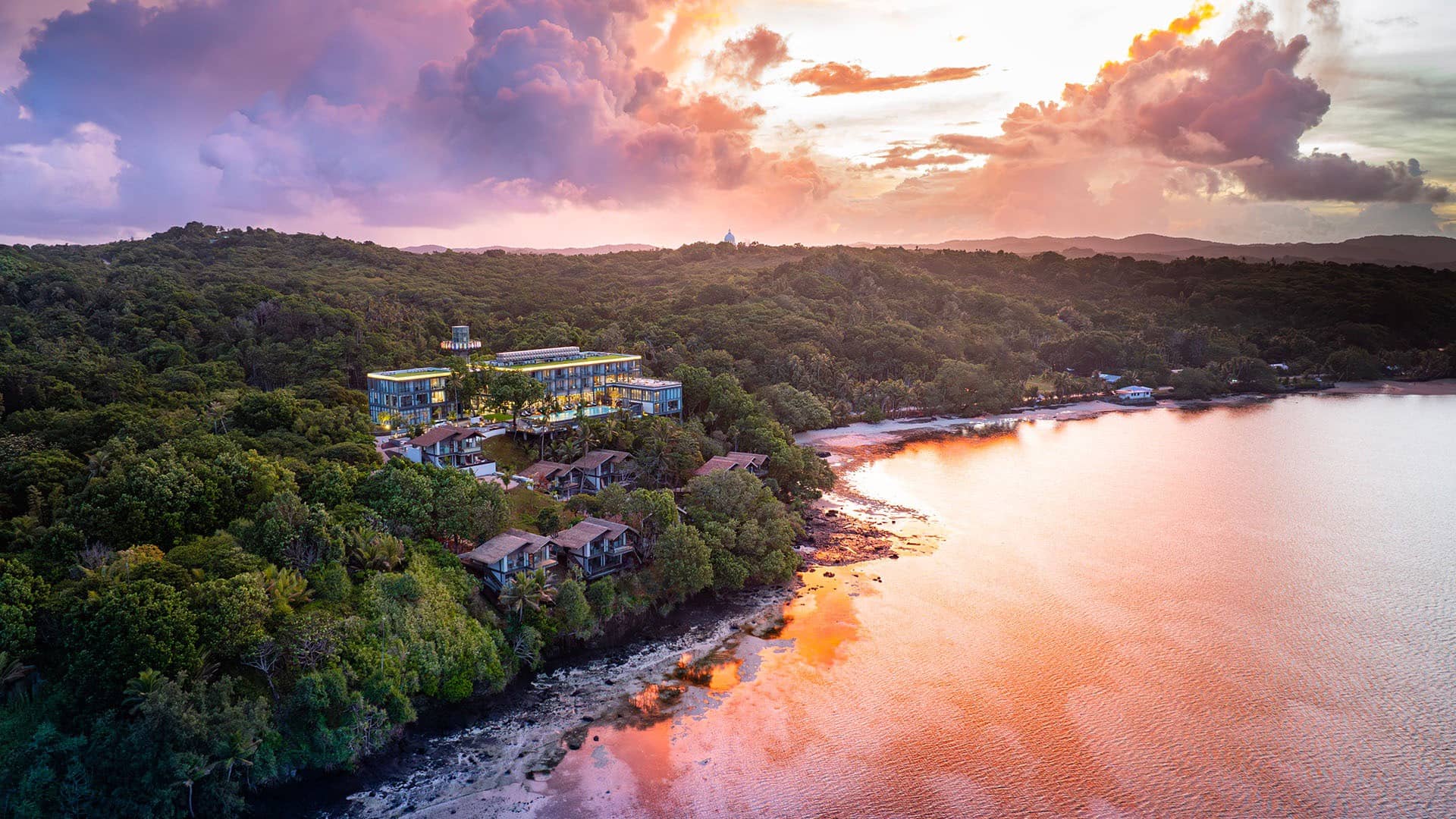 Dramatic sunset aerial view of tropical resort and villas nestled in jungle along tranquil bay with colorful sky