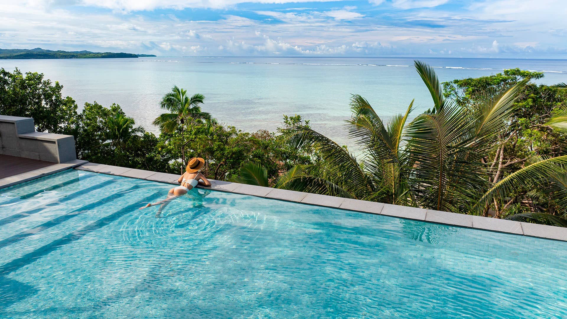 Woman relaxing in infinity pool with palm trees and serene ocean horizon under blue sky